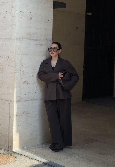 Mujer con gafas de sol oscuras y blazer negro y pantalones anchos sostiene un bolso negro trenzado, apoyada contra una pared de piedra en una zona exterior sombreada.