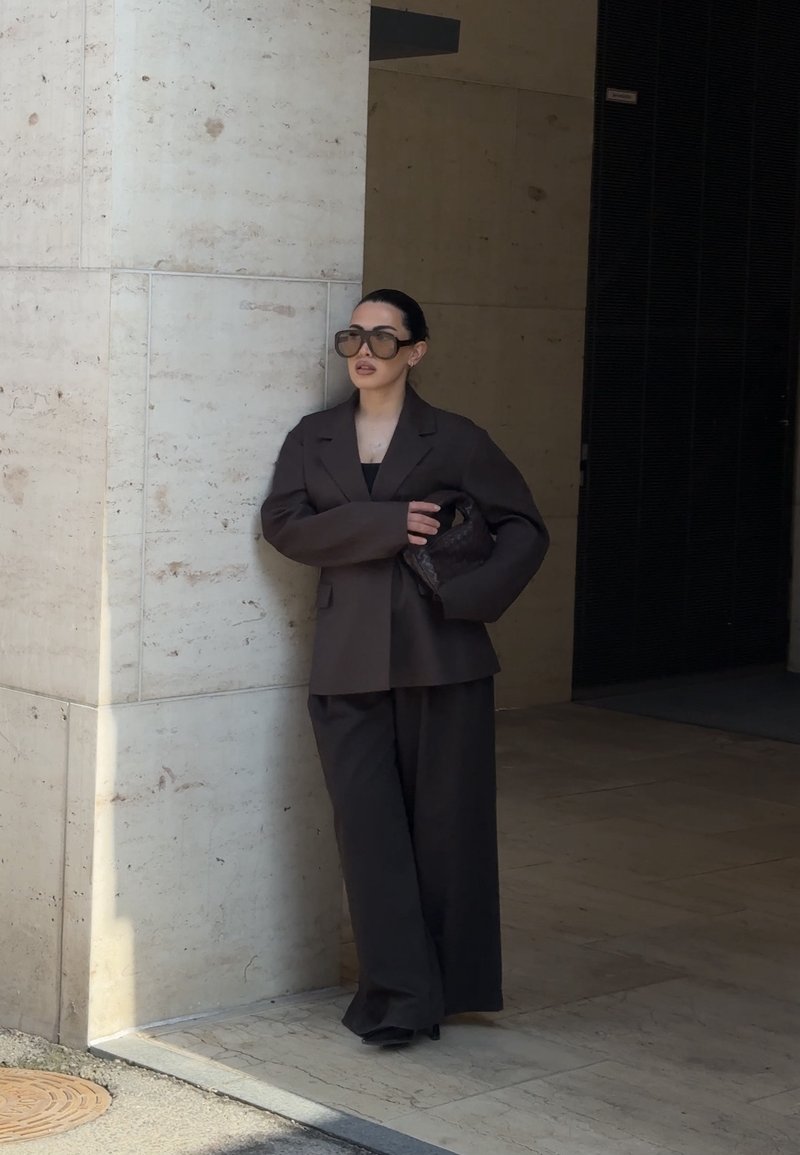 Woman with dark sunglasses and black blazer and wide pants holds a black braided handbag, leaning against a stone wall in shaded outdoor area.