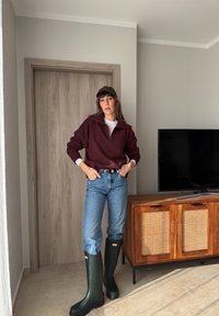 Femme portant un pull bordeaux, un jean bleu, une ceinture noire, des bottes de pluie noires et une casquette foncée, debout dans une pièce à côté d'un meuble TV en bois.