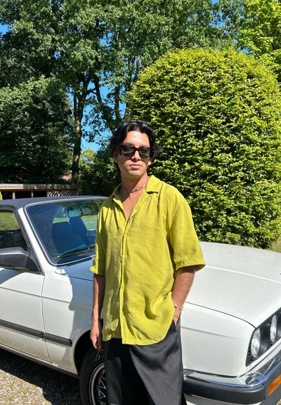 Camisa de lino verde claro de manga corta, pantalones negros, de pie junto a un coche plateado. Luz del sol, árboles al fondo, cielo azul visible.