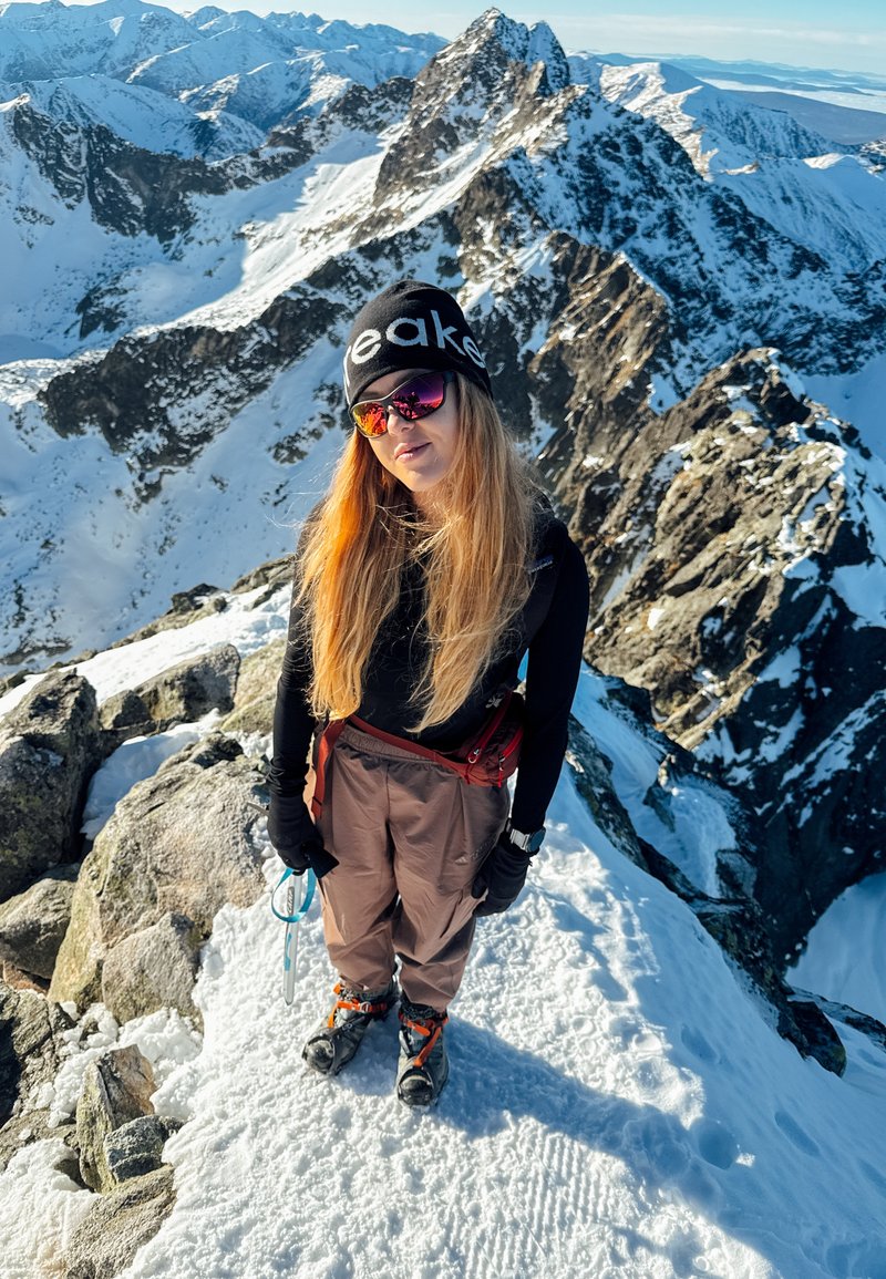 Un grimpeur portant un bonnet noir et des lunettes de soleil réfléchissantes, un haut noir à manches longues, un pantalon marron et des chaussures d'escalade, debout sur des rochers enneigés.