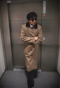 Homme portant un long manteau beige et des lunettes de soleil noires, debout les bras croisés devant des portes d'ascenseur fermées.