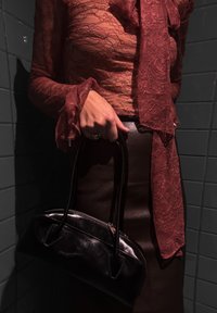 Fekete bőr kézitáska ívelt formával és két pánttal, amelyet egy kéz tart. Látható egy texturált bordó csipketop és egy barna bőrszoknya.