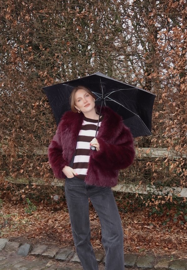 Femme en manteau de fourrure bordeaux et pull rayé tenant un parapluie noir debout sur un chemin couvert de feuilles, avec des arbres nus derrière elle.