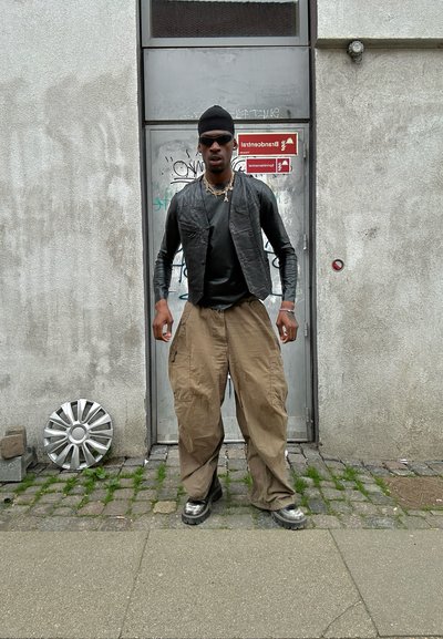 Individuo vistiendo un chaleco de cuero negro sobre una camiseta negra ajustada, con pantalones cargo beige sueltos y calzado oscuro, de pie frente a una pared.