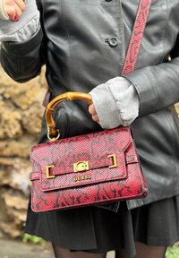 Czerwona torebka w wzór skóry węża z bambusowym uchwytem, złotymi elementami i zapięciem z logo marki. Noszona z czarną skórzaną kurtką i warstwowymi rękawami.