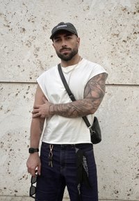 Weißes ärmelloses Hemd, dunkle Denim-Jeans mit schwarzen Akzenten, Umhängetasche. Auffällige Tätowierungen an den Armen, trägt eine schwarze Kappe und Sonnenbrille.
