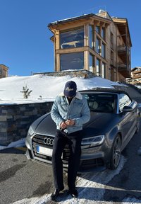 Homme en veste en jean et casquette debout à côté d'une Audi cabriolet grise garée près d'une colline enneigée et d'un chalet en bois moderne par une journée claire.