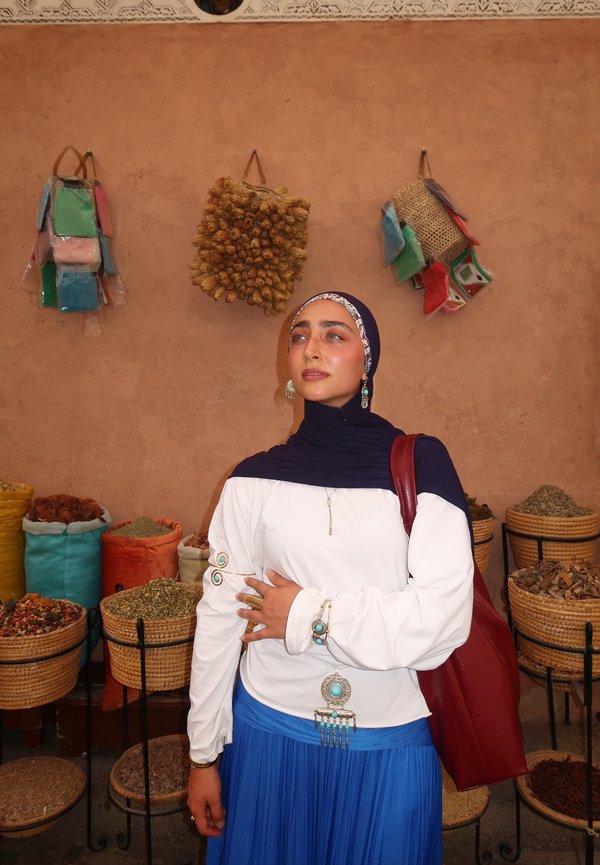 Woman wearing a navy hijab, white top, and blue skirt stands in front of baskets filled with spices and herbs in a market setting.