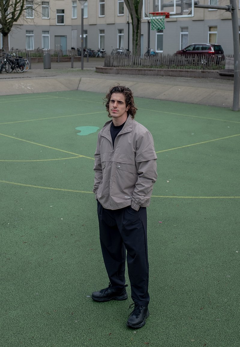 Šviesiai rudas švarkas su priekinėmis kišenėmis, juoda marškiniai, plačios juodos kelnės ir juodi sportbačiai, stovintis ant žaliai guminiu paviršiumi.