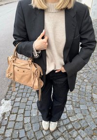 Άτομο με μπεζ πουλόβερ, μαύρο σακάκι με λεπτές ρίγες, μαύρο παντελόνι, λευκά αθλητικά παπούτσια και κρατώντας τσάντα από δέρμα σε ταμπ απόχρωση σε λιθόστρωτο δρόμο.