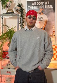 Sweat-shirt gris Nike avec logo ; porté sur une chemise à col. Porte un bandana rouge et des lunettes de soleil rondes, pantalon en denim foncé. Environnement intérieur.