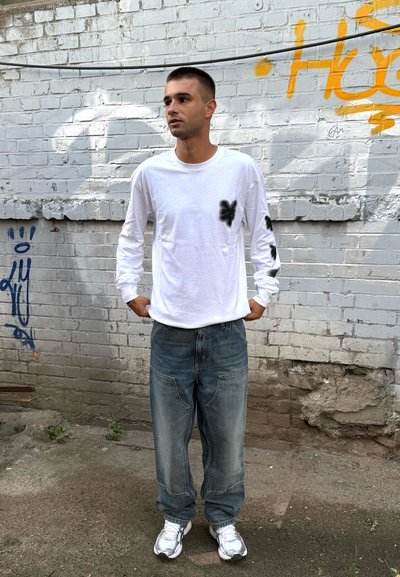 Camisa blanca de manga larga con acentos gráficos negros, combinada con jeans azules holgados y zapatillas plateadas, de pie contra una pared de ladrillo.