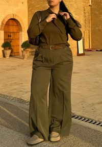 Femme en pull vert olive et pantalon large ajustant son col, debout à l'extérieur sur un pavé en pierre près de l'entrée d'un bâtiment historique.