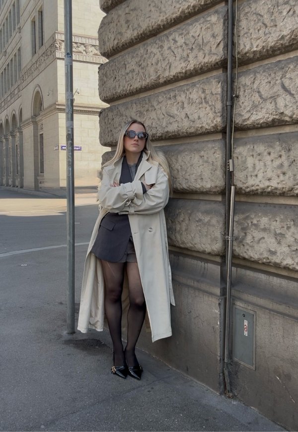 Mujer con abrigo largo beige, medias negras, gafas de sol y zapatos negros apoyada en la esquina de un edificio de piedra con textura en una calle de la ciudad.