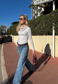 Haut gris à manches longues, écharpe beige clair, jean bleu taille haute et sac à main bordeaux. Marchant sur un trottoir avec un fond de haies.