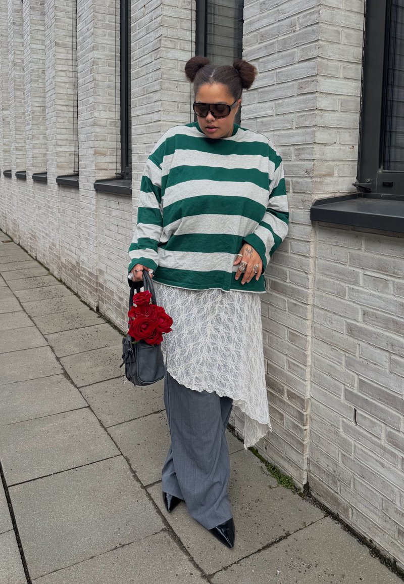 Person with two hair buns wearing sunglasses, green and white striped shirt, lace skirt overlay, gray pants, holding black bag with red roses.