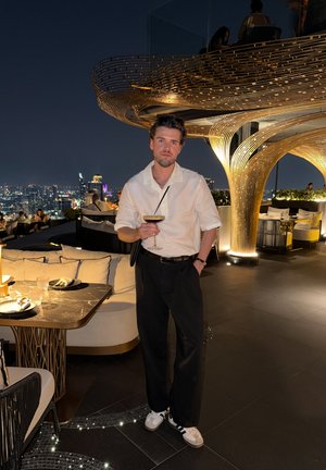 Hombre con camisa blanca y pantalones negros sosteniendo una copa de cóctel en un bar en la azotea iluminado con el horizonte de la ciudad de noche.
