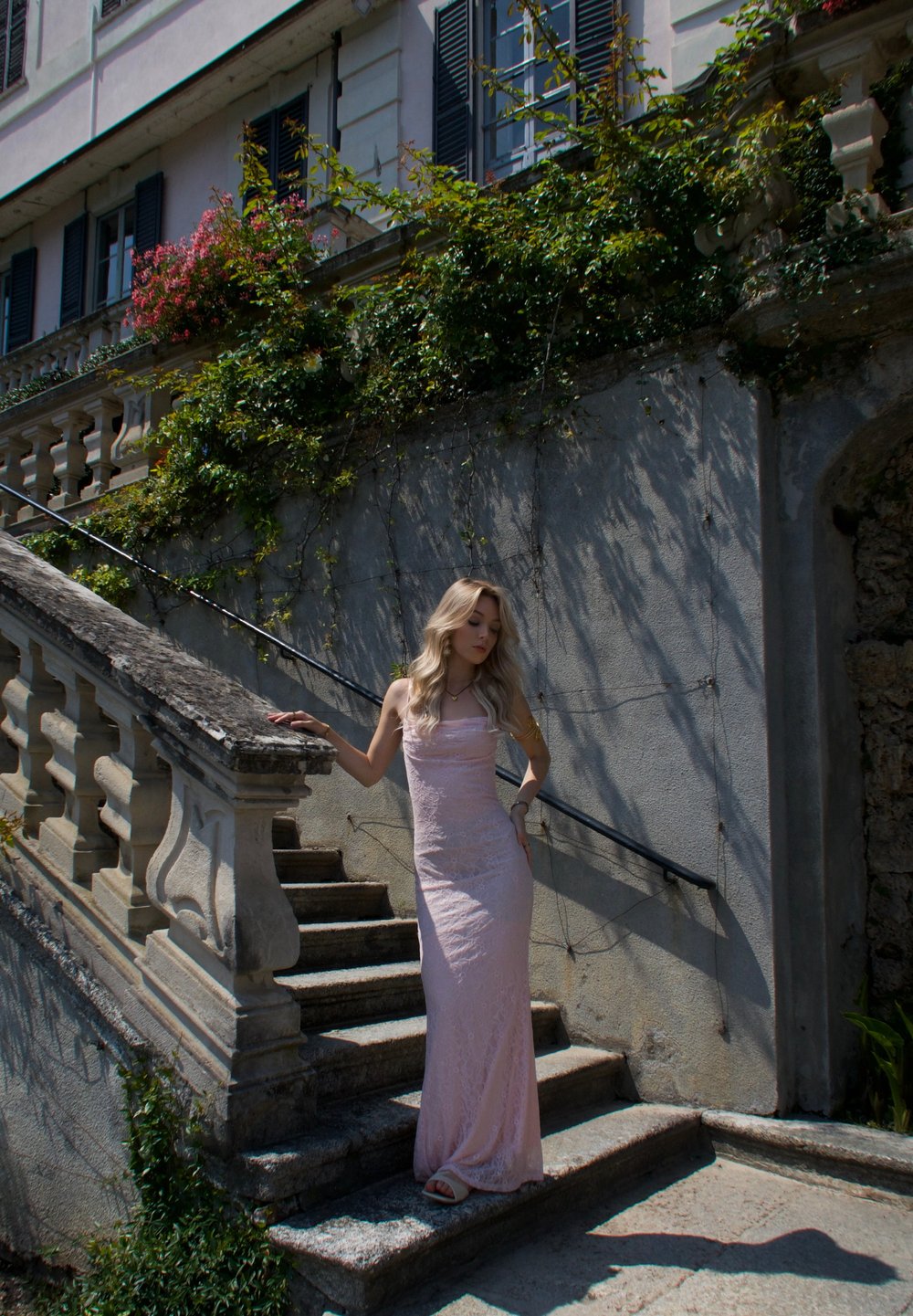 Abito lungo in pizzo rosa con design aderente, in piedi su scalini di pietra con parapetto; alberi e elementi architettonici sullo sfondo.