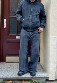 Homme portant une veste à capuche sombre, un jean gris ample et des chaussures noires épaisses, debout sur un seuil en pierre devant une porte marron.