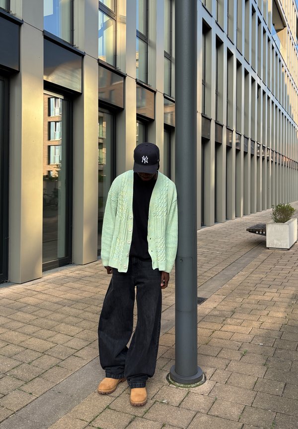 Cardigan lavorato a maglia verde chiaro con bottoni frontali, t-shirt nera, jeans neri larghi e stivali beige, in piedi accanto a un palo grigio in un contesto urbano.