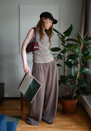Top sin mangas de canalé gris claro, pantalones marrones de gran tamaño, bolso burdeos, gorra negra, sosteniendo un libro. Planta verde y pared neutra de fondo.