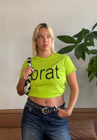 Svetlo limetno zelena kratka majica s črnim napisom "brat", v kombinaciji z modrimi kavbojkami in vzorčeno umetno krzno torbo s trakovi. Dekorativni pas s kovinskimi studom.