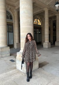 Φόρεμα με λεοπάρ σχέδιο, ψηλό γιακά, βολάν μανίκια και τριπλή σχεδίαση; συνδυασμένο με μαύρες μπότες μέχρι το γόνατο και μια μικρή τσάντα.