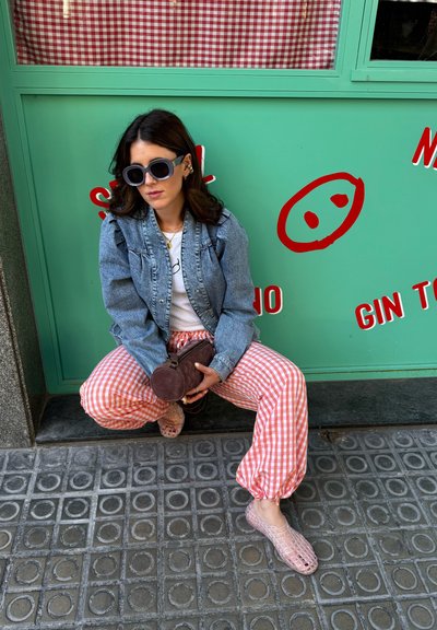 Chaqueta de mezclilla, camiseta blanca y pantalones de cuadros rosas. Zapatos claros y texturizados. Sosteniendo un bolso redondo pequeño y oscuro. El fondo es verde menta con acentos rojos.