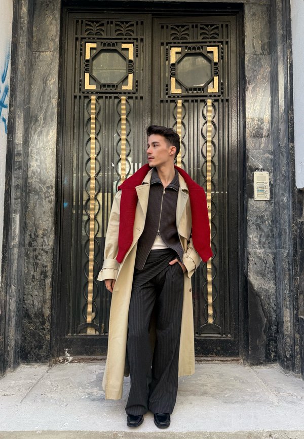 Jonge man met een beige trenchcoat en een rode trui over de schouders hangt, staat voor een sierlijke zwarte en gouden metalen deur.