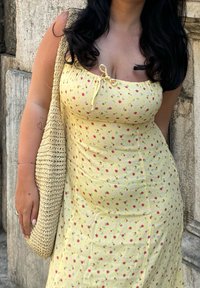 Κίτρινο φλοράλ φόρεμα με εφαρμοστή σχεδίαση, διακοσμημένο με μικρά ροζ και πράσινα μοτίβα, και φιόγκο στον λαιμό. Υφασμένος καλάθι από ψάθα.