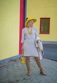 Γκρι υφασμάτινο φόρεμα με φουσκωτά μανίκια, κίτρινες αξεσουάρ και μπεζ μπότες μέχρι το γόνατο. Στηρίζει το σώμα της σε επιφάνεια από καλίβες πέτρες κοντά σε έναν κίτρινο τοίχο.