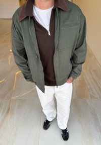 Chaqueta con cremallera color verde oliva y cuello de pana marrón, sobre un suéter marrón, combinada con pantalones blancos y zapatillas negras.