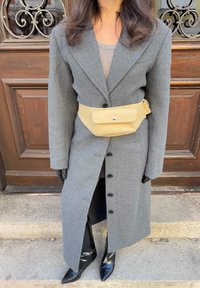 Manteau en laine gris à manches longues, fermé devant par des boutons, doté d'une ceinture-sac beige. Assorti à des bottes noires hautes et pointues pour un look structuré.