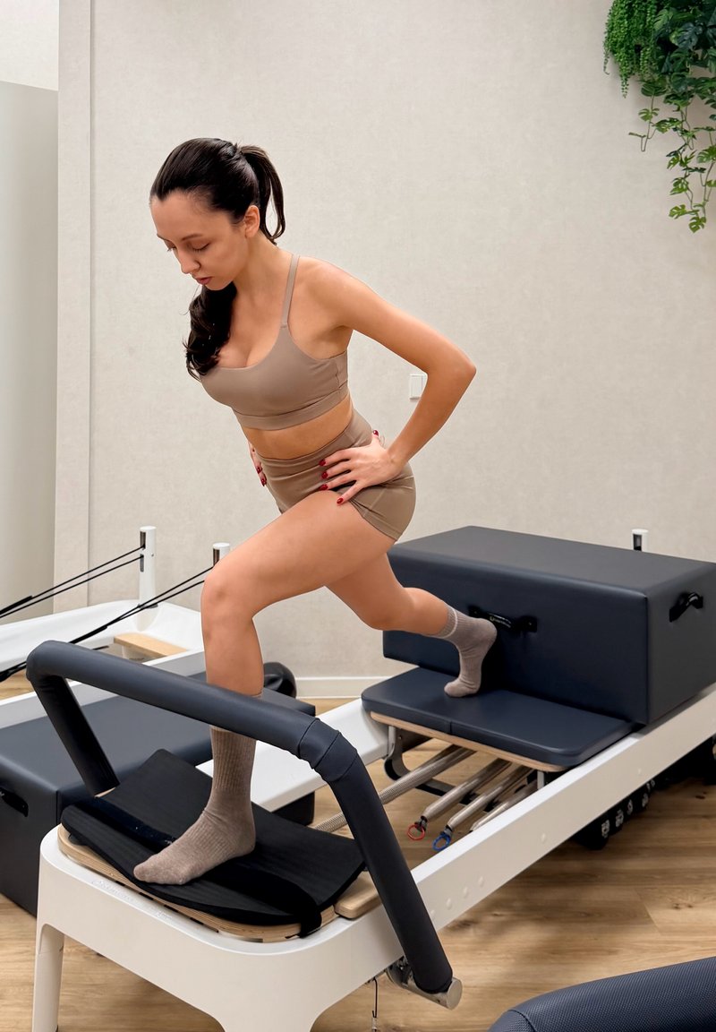 Pilates reformer i hvid og sort; polstrede overflader, justerbar fodstøtte, solid ramme. En bruger demonstrerer lunges.
