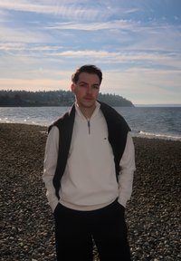 Jeune homme aux cheveux noirs portant un pull blanc et un pull noir, debout sur une plage rocheuse avec une mer calme et un rivage bordé d'arbres derrière lui.