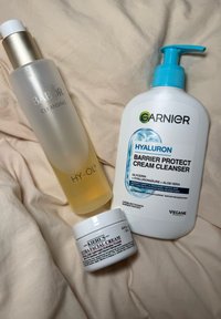 Tre hudplejeprodukter på beige stof: BABOR Cleansing Hy-Öl, Garnier Hyaluron Barrier Protect Cream Cleanser og Kiehl’s Ultra Facial Cream.