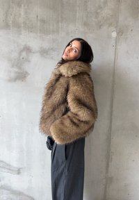 Barna műszőrme rövidített dzseki széles gallérral és túlméretezett ujjakkal, sötét, bő szabású nadrággal párosítva, beton háttér előtt.