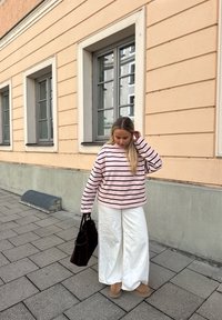 Pink og sort stribet oversized sweatshirt, hvide bukser med vide ben, brune støvler og en mørk taske, stående på en brostensbelagt fortov nær en bygning.