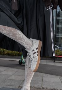 Baltas sneakers ar pelēkām zamša akcentiem, trim tumšām svītrām un gumijas zoli, apvienoti ar baltām zeķbiksēm un plisētu melnu svārku.
