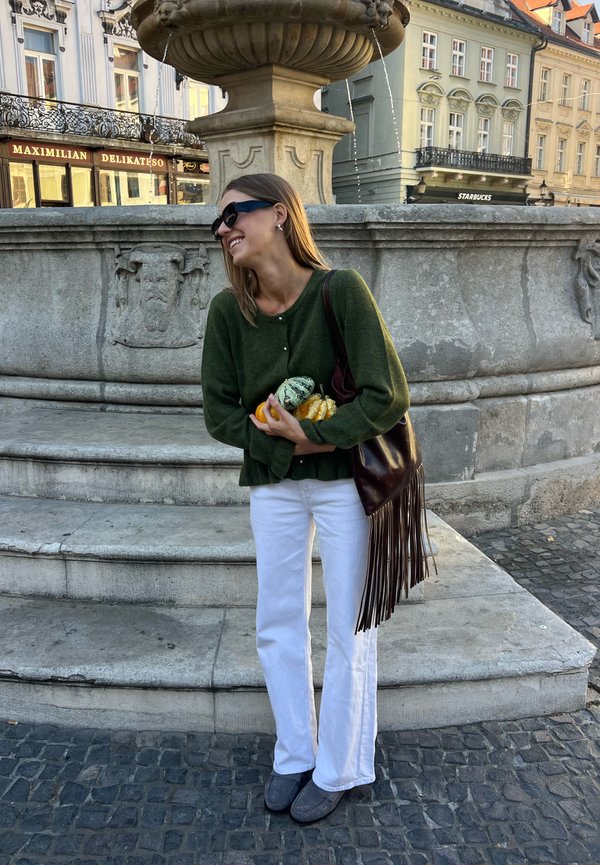 Una mujer con gafas de sol sonríe junto a una fuente de piedra en una plaza de la ciudad. Lleva flores en la mano y viste un jersey verde y unos pantalones blancos.