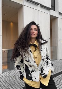 Mākslīgās kažokādas mētelis melnbaltā govs rakstā, apvienots ar spilgti dzeltenu pogājamās krekla krekliņu ar melnu loku apkaklīti. Redzami teksturēti auduma detaļas.