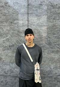 Siva majica dugih rukava s crnom beanie kapom; nosi teksturiranu bež torbu s šarenim printovima; u pozadini siva kamena površina.