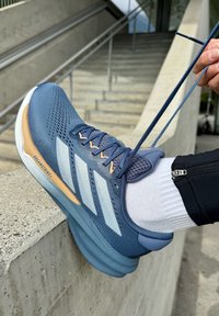 Blå sportssko med mesh-overdel og tre hvite striper, med polstret krage og en lys tan mellomsåle, som hviler på en kant.