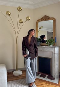 Blazer marron cintré avec fronces sur les côtés, pantalon large gris, et sac à main argenté ; intérieur moderne avec lampe sur pied dorée et miroir.