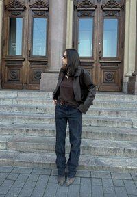 Σκούρος καφέ δερμάτινος μπουφάν πάνω από σκούρο καφέ τοπ, συνδυασμένο με μαύρα τζιν και γκρι μποτάκια με μυτερές μύτες, στέκεται σε πέτρινες σκάλες.