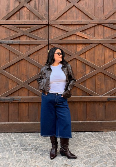 Mujer con gafas de sol, chaqueta de cuero marrón, camisa blanca, vaqueros anchos azules y botas marrones, de pie sobre adoquines frente a una puerta de madera.