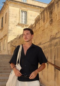 Zwart shirt met korte mouwen, witte shorts en een canvas tote bag. Houdt een zonnebril vast, staat op stenen trappen naast een gestructureerde beige muur.