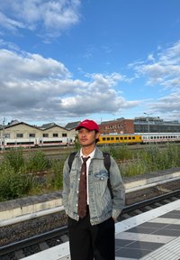 Mladý muž s červenou čiapkou, rifľovou bundou a kravatou stojí na nástupišti vlakovej stanice, za ním sú vlaky a priemyselné budovy.