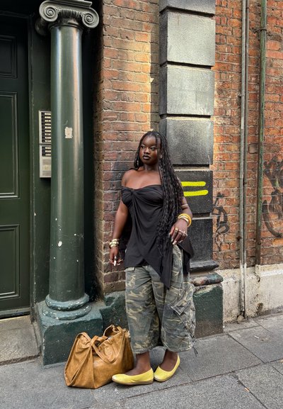 Top negro sin hombros, pantalones cargo de camuflaje, zapatos planos amarillos, accesorios dorados, bolso de cuero marrón, frente a una pared de ladrillos texturizada.